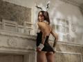 SophieKitty with tattoos, black bunny outfit, white cuffs, and bunny ears, showing her bare ass, looking back, standing in front of ornate white fireplace.