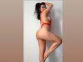 HeidyMillerr in red lace lingerie, side profile, showing her big ass, small tits, and raised leg, with black hair in a ponytail, against a white wall.