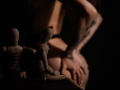 SophieKitty's tattooed arm covers her ass, hands resting on her butt. Two wooden artist mannequins are in the foreground, partially shadowed. Dark, moody lighting.