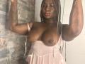 AkaneCox with dark skin, braided hair, red lipstick, large bare boobs, pink sheer dress, holding straps, tattoo on right arm, standing in a shower, looking at camera.