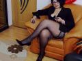 ClassybutNaughty with black hair, glasses, and a black blazer, sitting in an orange leather chair, wearing black stockings and heels, showing her tits and holding a remote.