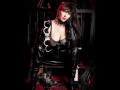 MaitresseElsa with big boobs, black leather outfit, cuffs, and choker, sitting on a chair, holding handcuffs, red light, and leather accessories on the floor.