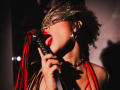 AliceJelanni with curly hair, red lipstick, and gold mask, licking a black and red flog while holding it with her hand, wearing a red halter and gold earrings.