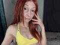 HannaCool with red hair, freckles, nose piercing, and lip piercing, wearing a yellow bra, touching her face, smiling slightly. Her tits are small, and she's looking at the camera.
