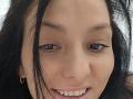 JoliDanielle's face, showing her brown eyes, black eyeliner, and white teeth with a slight smile. Her black hair is partially covering her face, and she's wearing gold earrings.