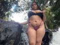 LolyMarce with big boobs in a black bra and purple thong, standing by a waterfall, holding her bra, water dripping on her tan skin, looking down at the camera.