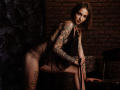 SophieKitty with tattoos, wearing sheer black lingerie, leaning on a chair, showing her ass, holding a black strap, brown bob, dark eyes, sexy, brick wall background.