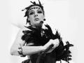 Black-and-white photo of MaitresseElsa with feathers, wearing a headpiece, bracelet, and holding a whip, looking down with a serious expression. Boobs partially covered by dark feathers.