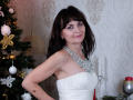 AngieGreen with dark brown hair, pink eyeshadow, white strapless dress, and silver necklace, standing by a decorated Christmas tree, showing her small, perky tits.