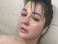 AlinaMilf with wet black hair, droopy eyelids, and glistening skin, water spraying on her face, pink lips slightly open, and small gold earrings visible in a shower.