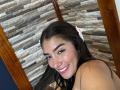 SharonKlart has long dark hair, a white hairclip, and is smiling with red lipstick, showing off her tits, in a selfie with wooden and stone wall background.