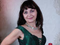 AngieGreen with dark brown hair, green satin dress, lace-up cleavage, purple eyeshadow, silver necklace, smiling, medium tits, fair skin, standing, slight red light on left.