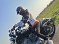 FrancaiseAnnaDom with black helmet, reflective visor, white-and-black jacket, orange shirt, black pants, and white sneakers, riding a black motorcycle on a sunny road.
