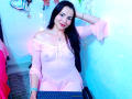 EmelySofia with dark hair, pink sheer dress, visible nipples, sitting, hand on head, smiling, keyboard in front, blue lighting, pink wall, shiny heart decoration, bracelet.