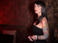 MissVonTease with black hair, red lipstick, and a strapless black dress, holding a remote. She's got colorful tattoos on her right arm and a bracelet on her left wrist.