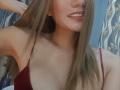 DianaShane with long brown hair, smiling, wearing a red spaghetti strap top, showing her chest, small earrings, and her hand in her hair, in a selfie.