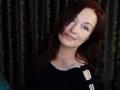 MarleneDavies has pale skin, blue eyes, and dark red hair, wearing a black top with white straps, smiling slightly. Her tits are small and she looks cute.