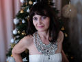 AngieGreen with dark brown hair, wearing a white strapless dress and a chunky silver necklace, standing in front of a decorated Christmas tree, smiling slightly. Boobs visible.
