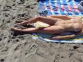 LaFrancaise is lying naked on a beach towel with yellow and white stripes, legs spread, exposing her pussy, small tits, and shaved pubic area, wearing sandals on the sand.