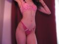 SweetClaraa has pink lace bra and matching pink panties with a white bow, standing with one arm raised, showing a slim, toned body and small, perky tits.