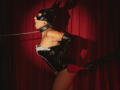 JuliaNewell in black leather mask, corset, and gloves, chained, with hands behind back, red curtains, and black-gloved hand holding red paddle, mouth open, eyes closed.
