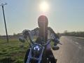 FrancaiseAnnaDom on a motorcycle, sun in back, wearing a red helmet and black/white jacket, gripping handlebars, road ahead, sun flaring, clear sky, power lines on left.