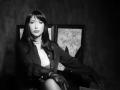 Black-and-white photo of MissVonTease with long dark hair, wearing a buttoned-up jacket, exposing her tits, gloved hands, and a necklace, sitting against framed art.