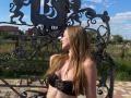 ArielleFiorello, with long brown hair, wearing a black strapless top with a floral gem, standing in front of an ornate metal gate, blue sky, and green grass.