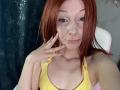 HannaCool with red hair, freckles, nose ring, and lip piercing, wearing a yellow tank top, touching her cheek, showing a slight smile. Her tits are small and her ass is not visible.