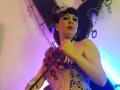 MaitresseElsa with short black hair, wearing a large black hat with metal rings, holding a black rod, dressed in a pink lace corset with roses, and a wrist bracelet.