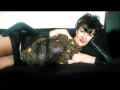 MaitresseElsa laying on a black couch, wearing a black lace dress with gold patterns, black leather gloves, and short black hair, showing her bare thighs and a sexy smile.