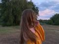 ArielleFiorello with long brown hair, wearing an off-shoulder yellow top, standing in a field, back to the camera, right hand touching her hair.