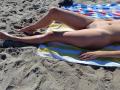 LaFrancaise's nude, tan-skinned body is lying on a striped towel at the beach, legs spread, displaying her clean-shaven, exposed pussy and a ring on her right hand.