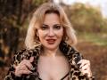 NatalyFoxy69 with blonde hair, wearing a leopard fur coat, holding it open to show her cleavage, smiling with red lipstick, and wearing gold earrings.