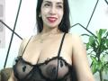 KatyKatt with dark hair, red lipstick, and smile, wearing sheer black lingerie that reveals her large, natural tits with visible nipples. Potted plant in background.