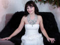 AngieGreen with pale skin, dark brown hair, smiling, wearing a strapless white dress and a glittery necklace, sitting on a black fur-covered sofa, arms resting on her thighs.