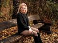 NatalyFoxy69 sits on a wooden bench, wearing a black sweater, black tights, and black boots, with blonde hair and red lipstick, in a forest with fallen leaves.