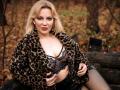 NatalyFoxy69 with blonde hair, leopard fur jacket, black lace bra showing her tits, black leggings, and dangling earrings, sitting on a log in a forest.