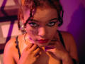 AliceJelanni with curly red-brown hair, wearing black lace, holding her tits, looking up with green eyes, red lipstick, and gold earrings, under purple and orange lighting.