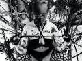 AmySpider, tattooed, wearing glasses, black strapless top, fishnet stockings, holding rope, nose ring, sitting among branches, black-and-white photo, visible tattoos, crouching, looking down, wearing fingerless gloves.