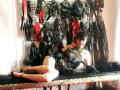 MaitresseElsa lies on a black table, wearing a black lace corset, holding a black leather whip, with various BDSM gear hanging in the background, and black heels on.