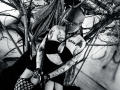 AmySpider with tattoos, bald with glasses, black bra, fishnet stockings, holding a whip, arms raised, dark cape, visible breasts, tattooed arms, and legs, in a black-and-white photo.