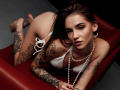 SophieKitty, with tattoos, in a white bikini, pearl necklace, on a red leather chaise, looking at the camera, showing her ass, long brown hair, and pierced ears.