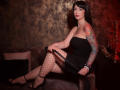 MissVonTease with black dress, polka-dot tights, black heels, and a tattooed arm, sitting on a brown couch, holding a black whip, red light on the right.