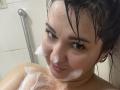 AlinaMilf with wet black hair, soap suds on her face and chest, looking at the camera with a slight smile, brown eyes, and dark eyebrows in a shower.