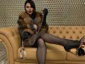 CrystalFetish with dark hair, red lipstick, fur coat, black gloves, fishnet stockings, high heels, and a silver chain holding handcuffs, sitting on a tan couch.