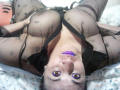 ZoeHotXX is lying on her back with her big tits showing through a sheer black dress, purple lipstick, and brown eyes looking up. She's wearing a black bra too.