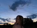 ArielleFiorello with long brown hair, small tits, and a shy smile, looking over her shoulder at the camera during a dark blue sunset, with her hand near her mouth.