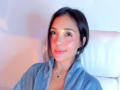 Linsay Colemann, avec ses cheveux foncés, sa robe bleue, son sourire discret, un léger blush rosé sur ses joues, un petit collier, une chaise blanche, un éclairage doux, un léger reflet de lumière de l'appareil photo sur son front..