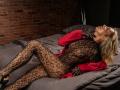 LettieSweet is lying on a bed, wearing a black lace bodysuit and red shirt, with her eyes closed and head tilted back, showing off her snake-patterned booty.
